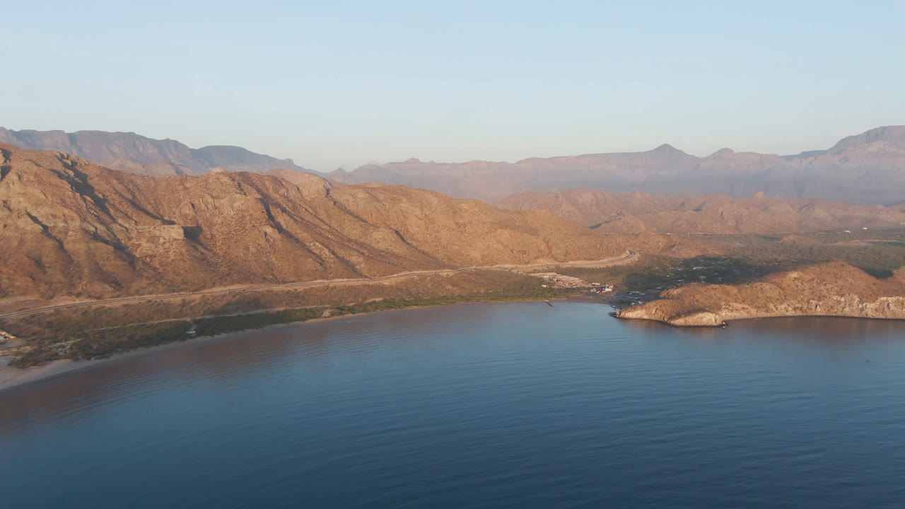 Aerial frame over Danzante Bay, Loreto, with open blue water in front, a small rocky island to the right of center, tan mountains behind it, and no visible people.