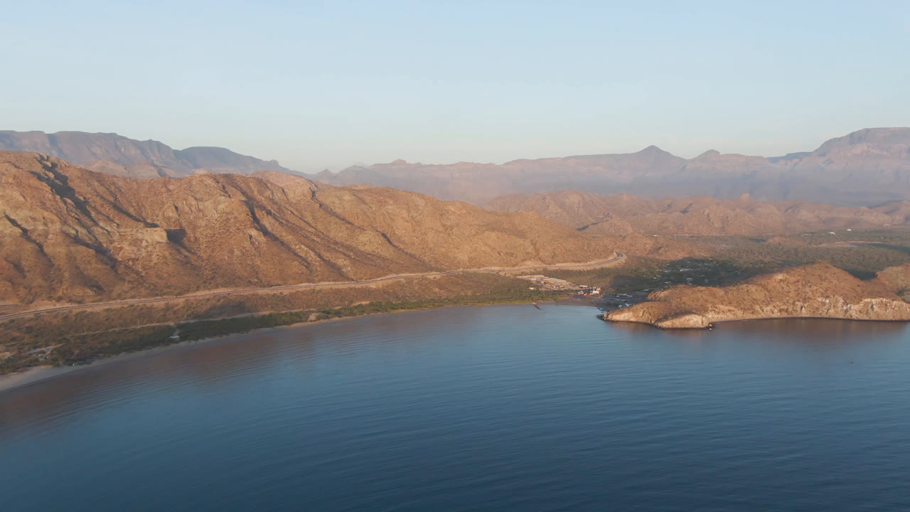 Second-frame aerial view of Danzante Bay showing smooth blue water, the island slightly right of center, a pale shoreline, layered desert mountains, and no visible people.