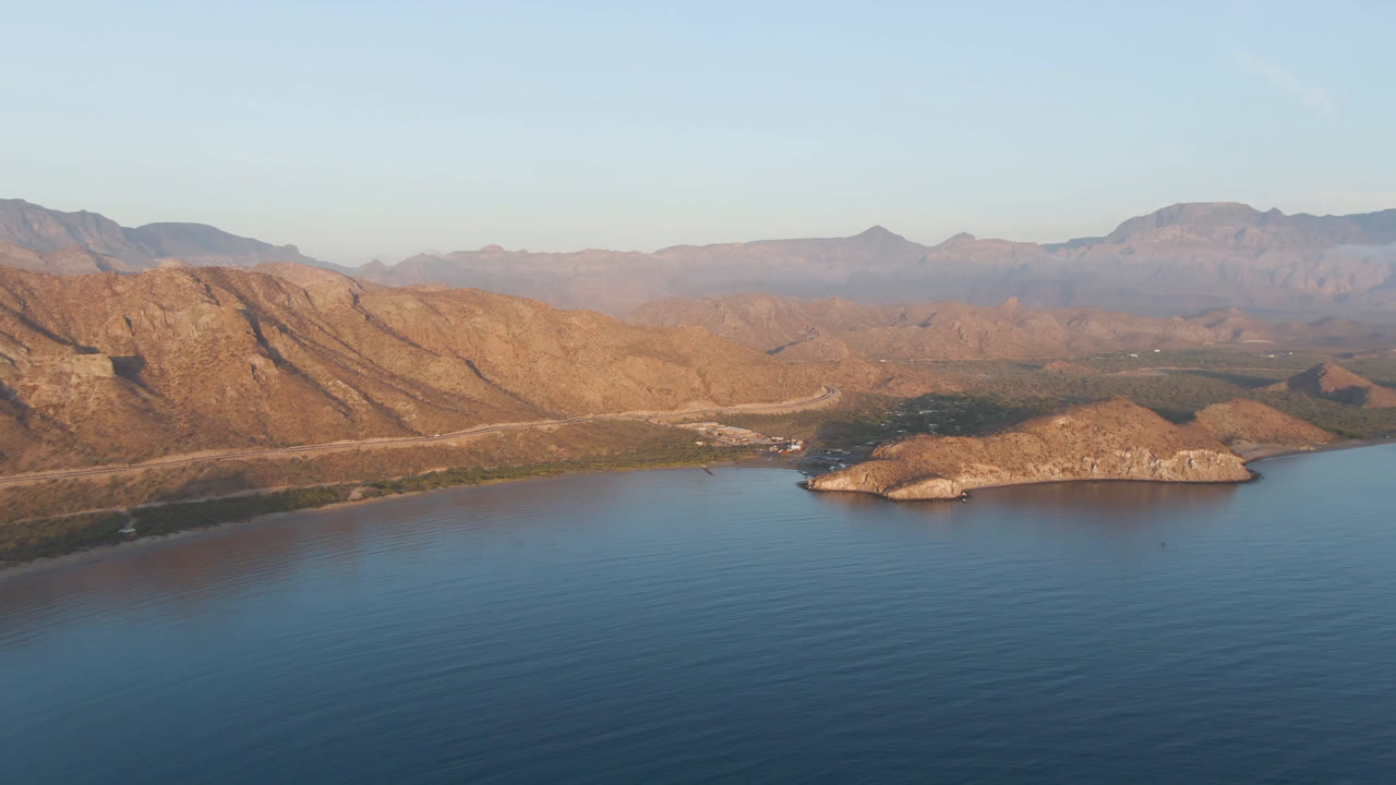 Opening aerial frame over Danzante Bay Loreto, Baja California Sur, Mexico, with blue water in front, a small rocky island near center, tan mountain ridges behind it, and no visible people.