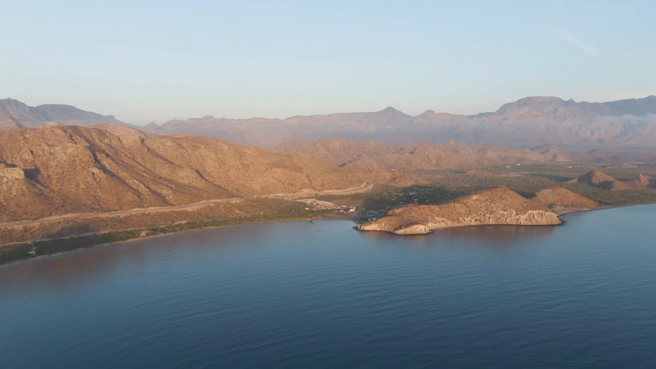 At two seconds, the high drone view shows Danzante Bay water in the foreground, a low rocky island in the midground, desert shoreline behind it, mountains across the horizon, and no visible people.