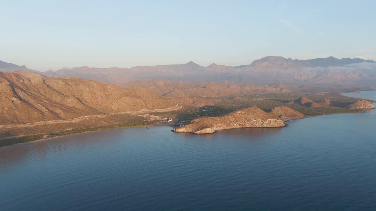 Aerial view of Danzante Bay with open blue water filling the foreground, tan ridges crossing the midground, small rocky islands near shore, distant mountains under pale sky, and no visible people.