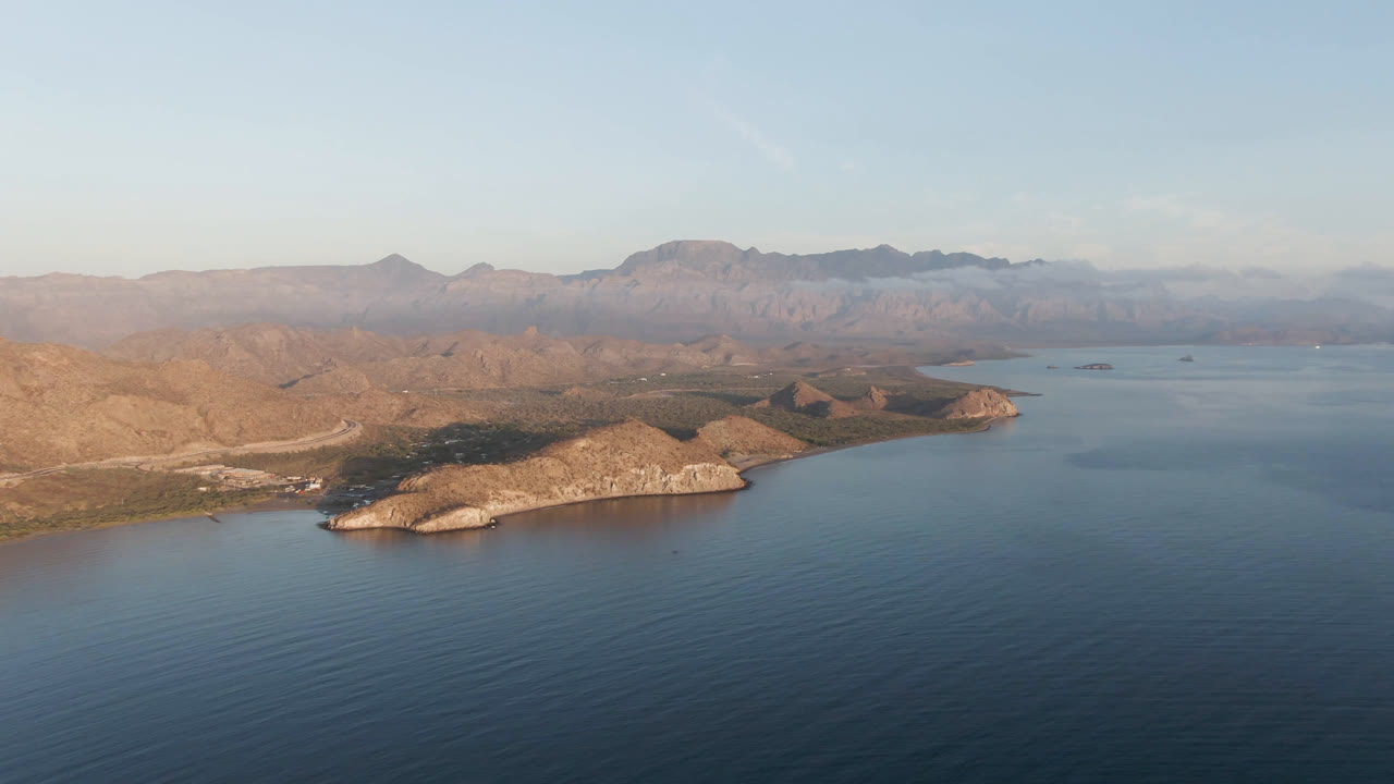 Aerial view over Danzante Bay in Loreto, Baja California Sur, with blue water in the foreground, a pale rocky point along the shore, hazy mountain ridges behind it, and no visible people.