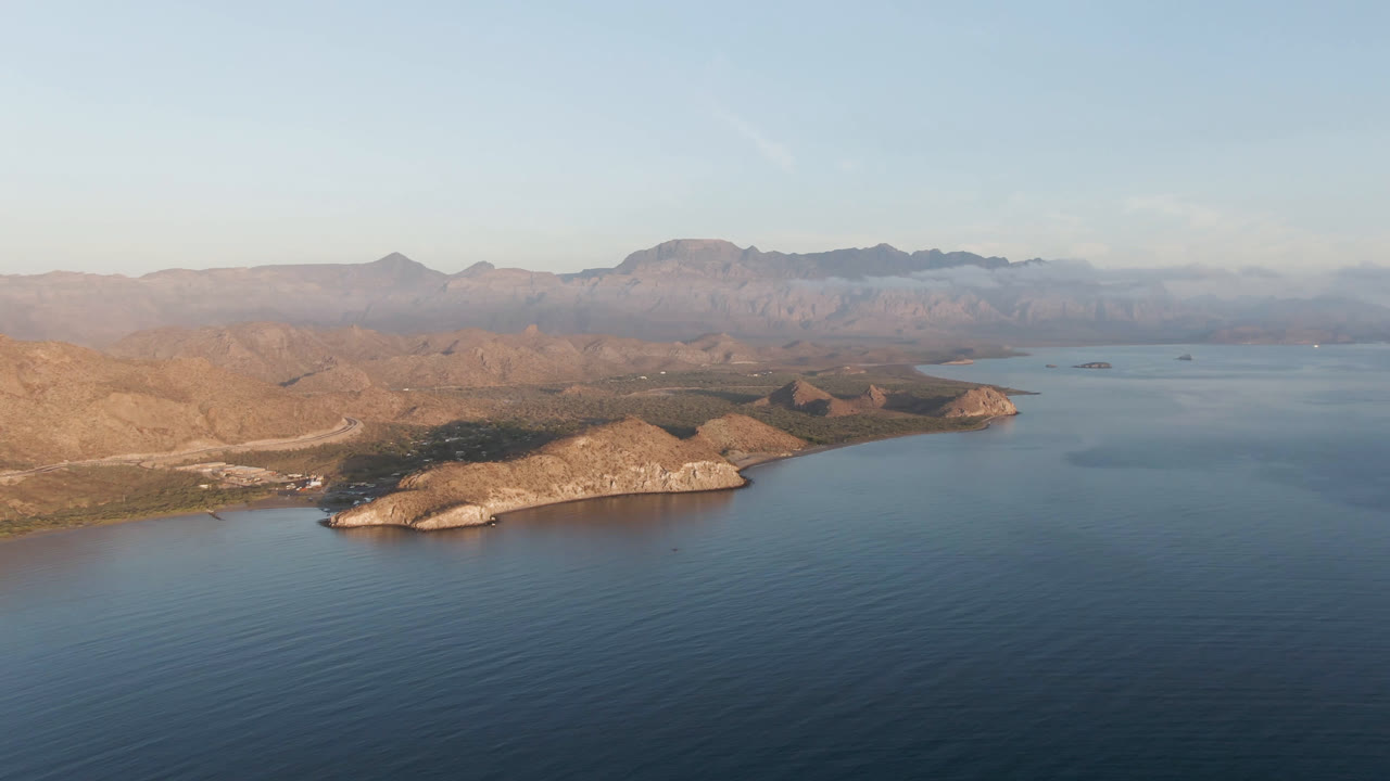 Wide aerial frame of Danzante Bay showing calm blue water below, a pale headland and sandy edge at center, dry hills beyond, distant mountains, and no visible people.