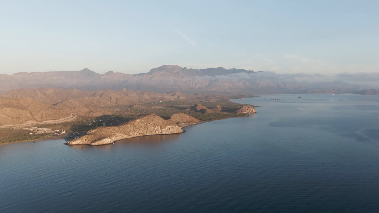 Aerial frame over Danzante Bay Loreto, Baja California Sur, with blue water in the foreground, a tan rocky point at left, distant mountains, and no visible people.