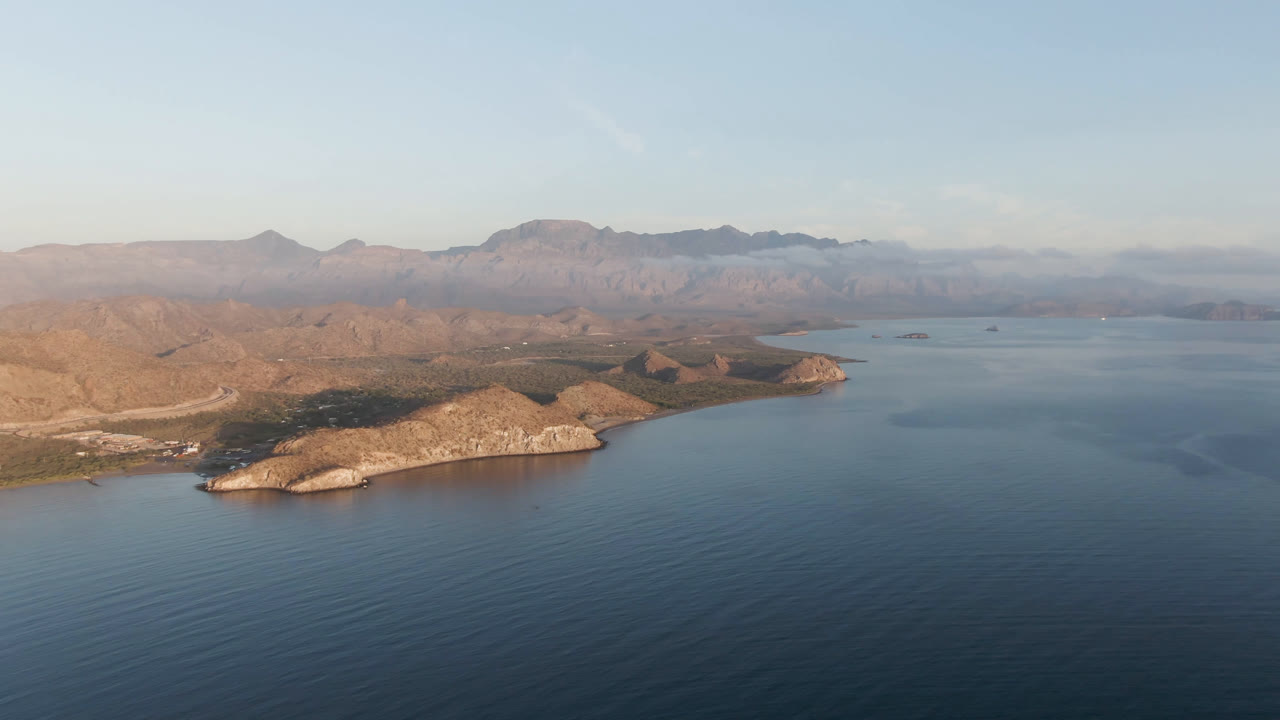 Aerial view of Danzante Bay Loreto, Baja California Sur, with the rocky peninsula slightly left of center, dark blue water below, pale sky above, and no visible people.