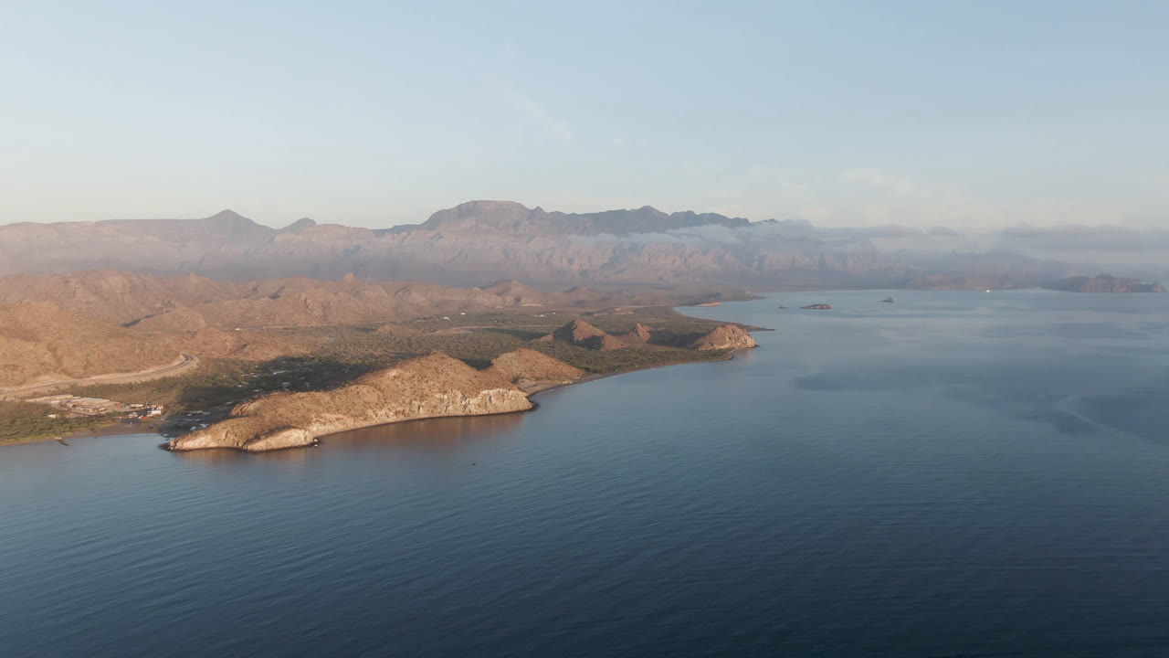Aerial coastal view with open water, low rocky land, and hazy mountains on the horizon.
