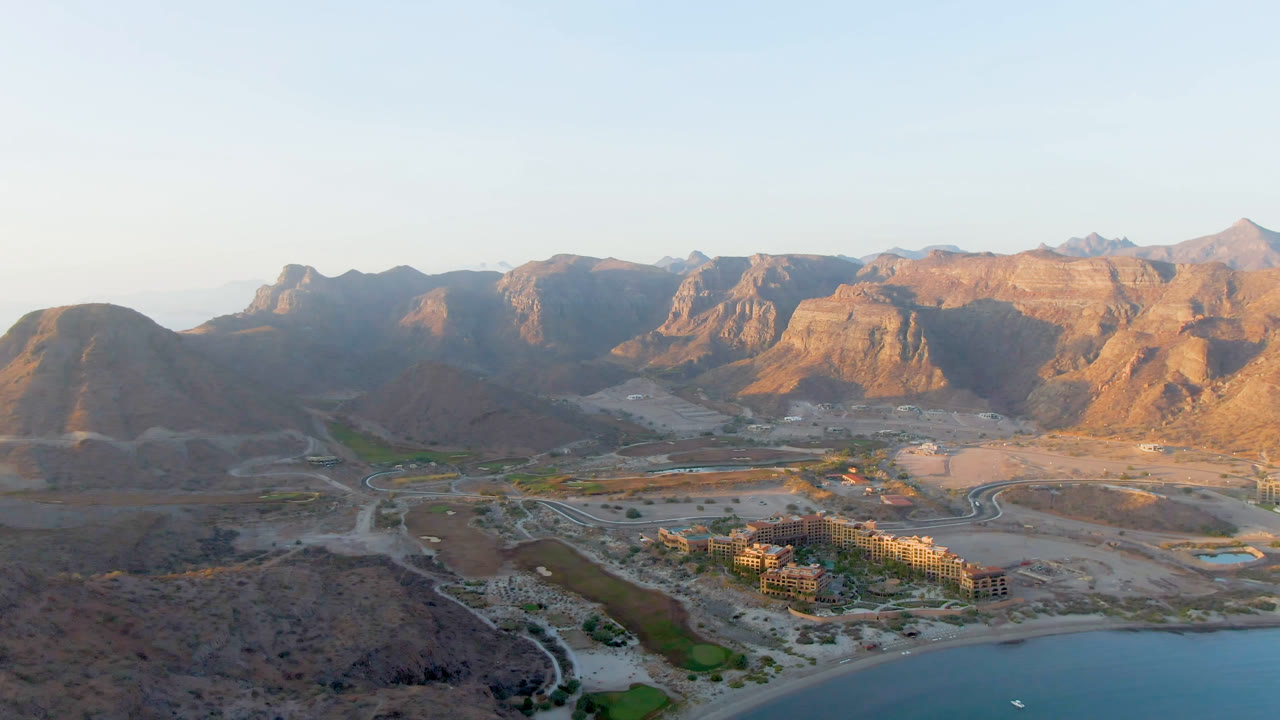 Aerial view over Danzante Bay in Loreto, Baja California Sur, with blue water along the lower edge, a building complex and roads in the valley, rocky mountains beyond, and no visible people.