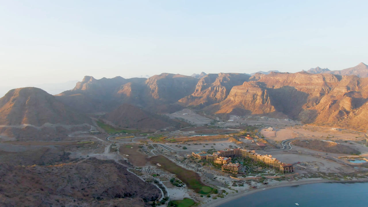 Wide aerial frame of Danzante Bay with shoreline water at bottom, a compact building complex near palms, pale desert roads, sunlit mountain ridges, and no visible people.