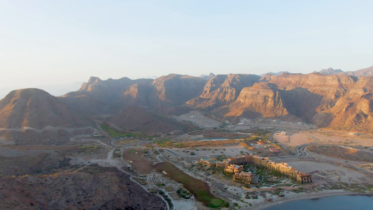 Opening aerial frame over Danzante Bay Loreto, Baja California Sur, showing tan mountains, winding roads, green landscaping, low buildings, water at the bottom edge, and no visible people.