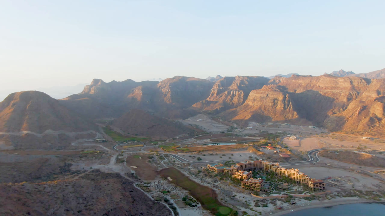 Second aerial frame of Danzante Bay Loreto with rugged sunlit mountains behind a dry basin, curving roads, a compact building cluster, landscaped green strips, and no visible people.