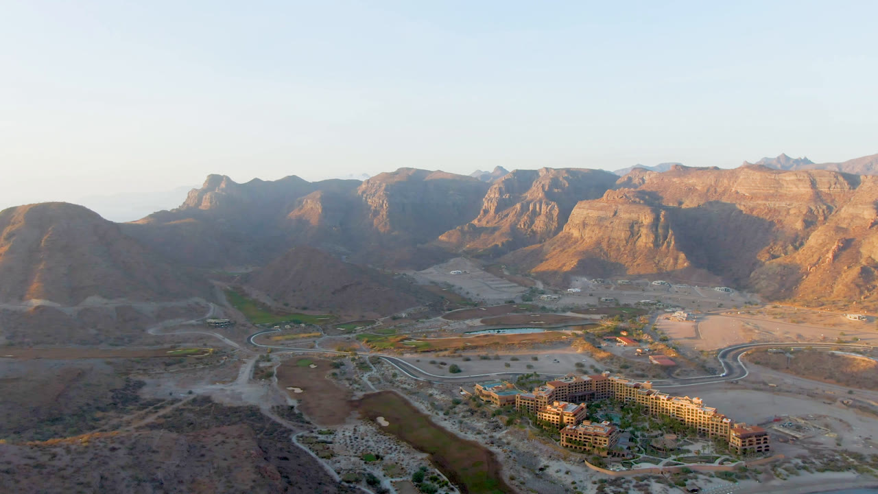 Opening frame of Danzante Bay, Loreto, with resort buildings low in frame, a green water channel, winding roads, rugged desert mountains, pale blue sky, and no visible people.