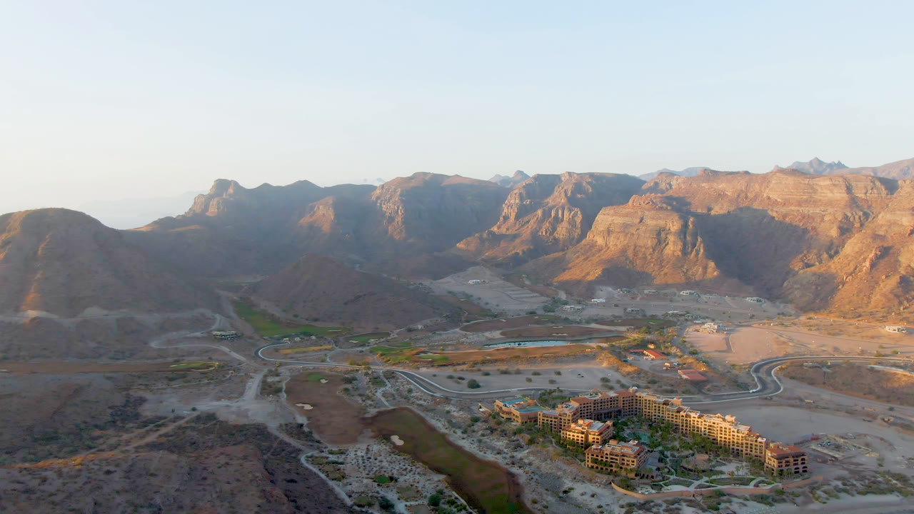Second frame over Danzante Bay, Loreto, showing the resort complex slightly lower in the view, curving roads through dry terrain, a narrow water channel, sunlit mountains, and no visible people.