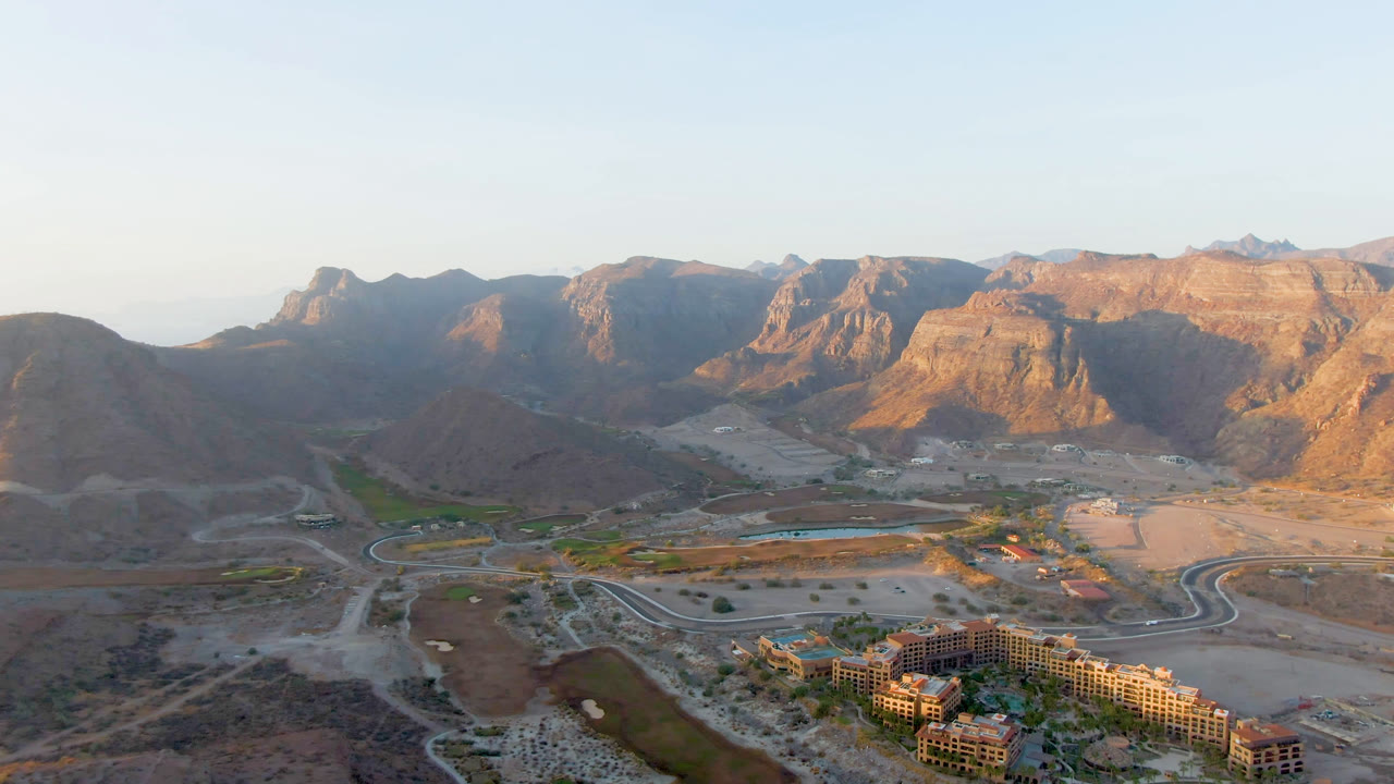 Drone view in Danzante Bay Loreto with the lower buildings slightly more central, a pale road network through the dry valley, green palms near water, and no visible people.