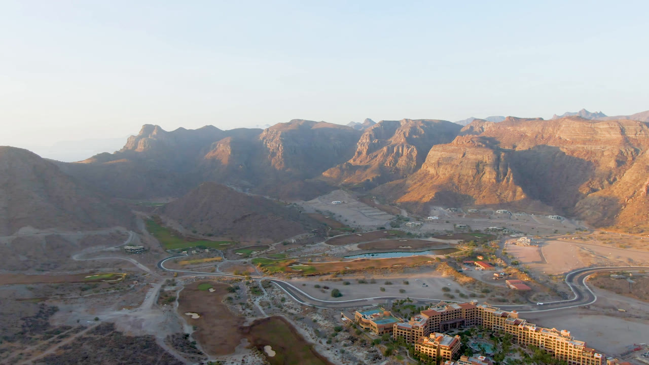 Wide aerial frame over Danzante Bay Loreto showing resort rooftops, palm landscaping, curved roads, and tan mountains in warm light with no visible people.