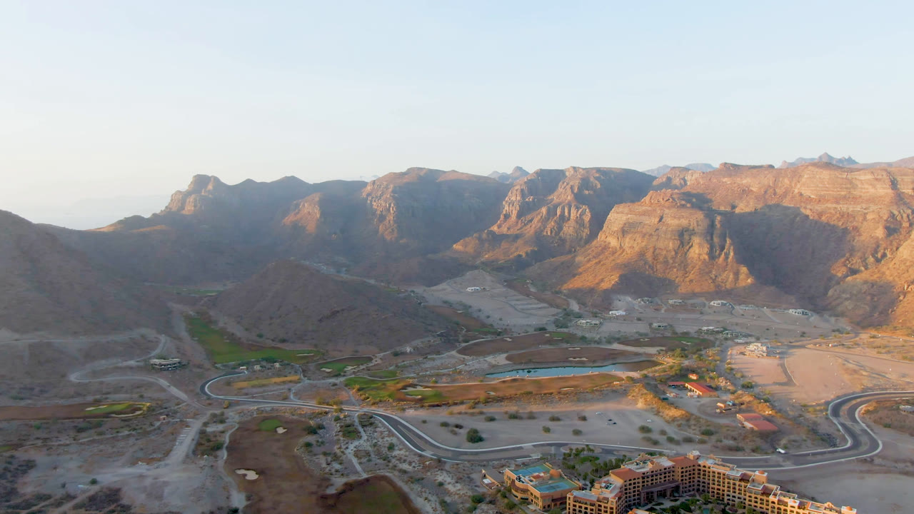 Wide aerial frame over Danzante Bay Loreto, Baja California Sur, Mexico, showing brown mountain walls, pale desert roads, resort grounds, blue pool water, and no visible people.