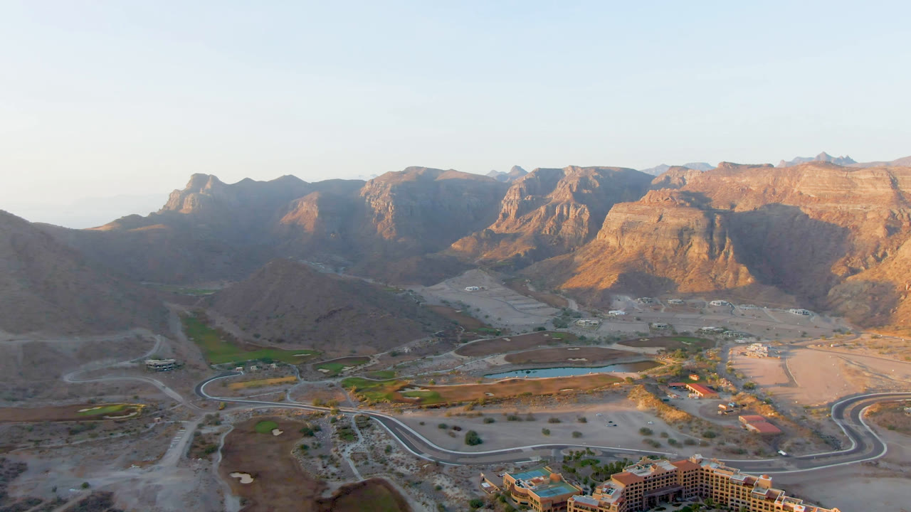 Aerial view in Danzante Bay Loreto with the resort edge low in frame, curved roads crossing desert flats, green landscaped patches, and sunlit mountains behind, with no visible people.