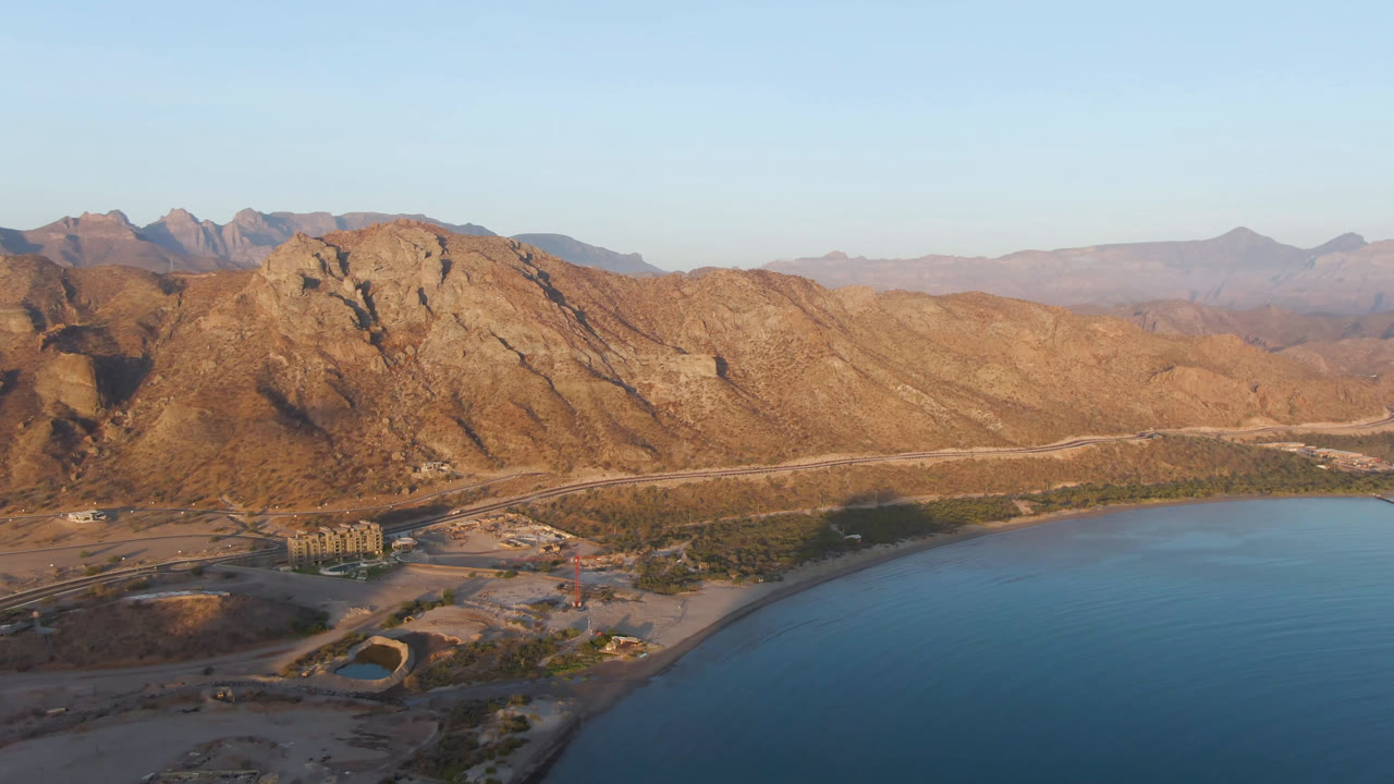 Wide aerial view of Danzante Bay in Loreto, Baja California Sur, with tan mountains, a curving blue bay, a shoreline road, and no visible people.