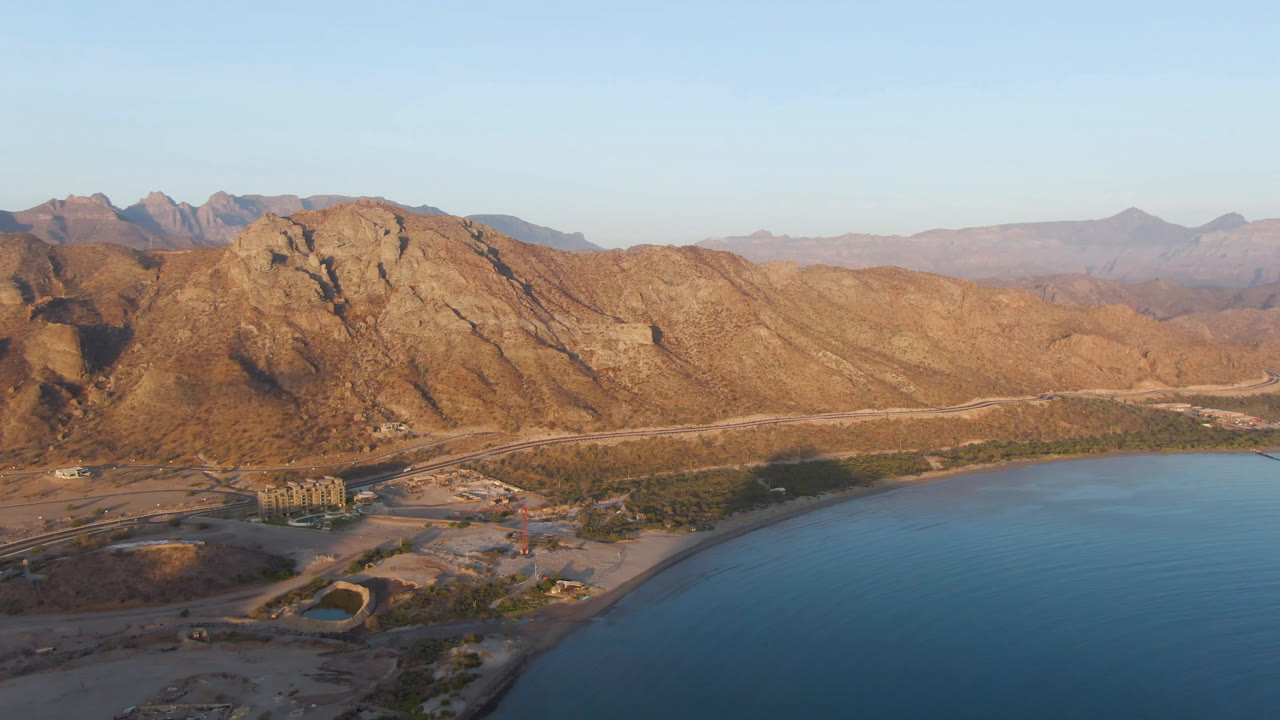 Aerial frame over Danzante Bay with rocky hills catching warm light, red-brown shoreline below, calm blue water at right, and no visible people.