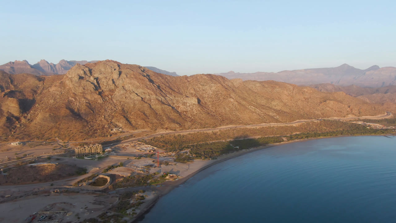 Opening aerial frame of Danzante Bay in Loreto, Baja California Sur, with tan mountains, blue water, a pale shoreline, a winding road, and no visible people.