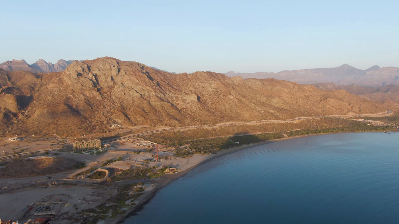 Aerial view over Danzante Bay Loreto with rocky tan mountains, blue water in the foreground, a curving road near the shoreline, and no visible people.