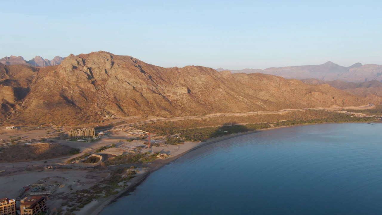 Opening aerial frame over Danzante Bay Loreto, Mexico, with blue water at lower right, shoreline roads and scattered buildings at lower left, rugged mountains in back, and no visible people.
