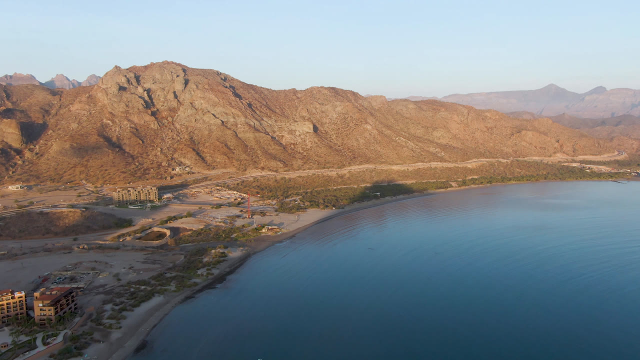 Aerial frame over Danzante Bay Loreto showing blue water at lower right, a curved shoreline with roads and low buildings, rugged brown mountains beyond, and no visible people.