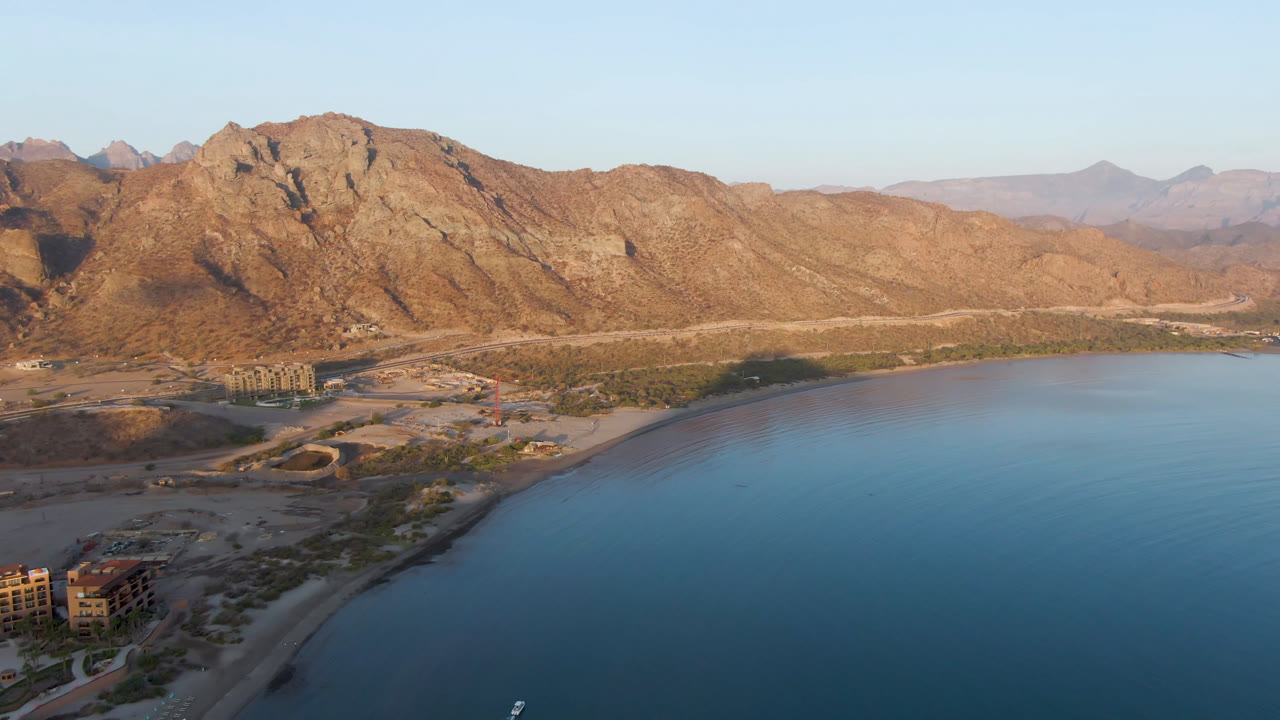 Drone frame over Danzante Bay Loreto with calm blue water occupying the lower half, sandy shore and coastal road in the midground, mountains in warm light, and no visible people.