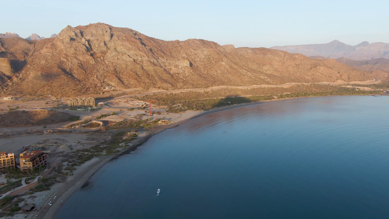 Aerial view of bay water, curved beach, desert mountains, and a small boat offshore.