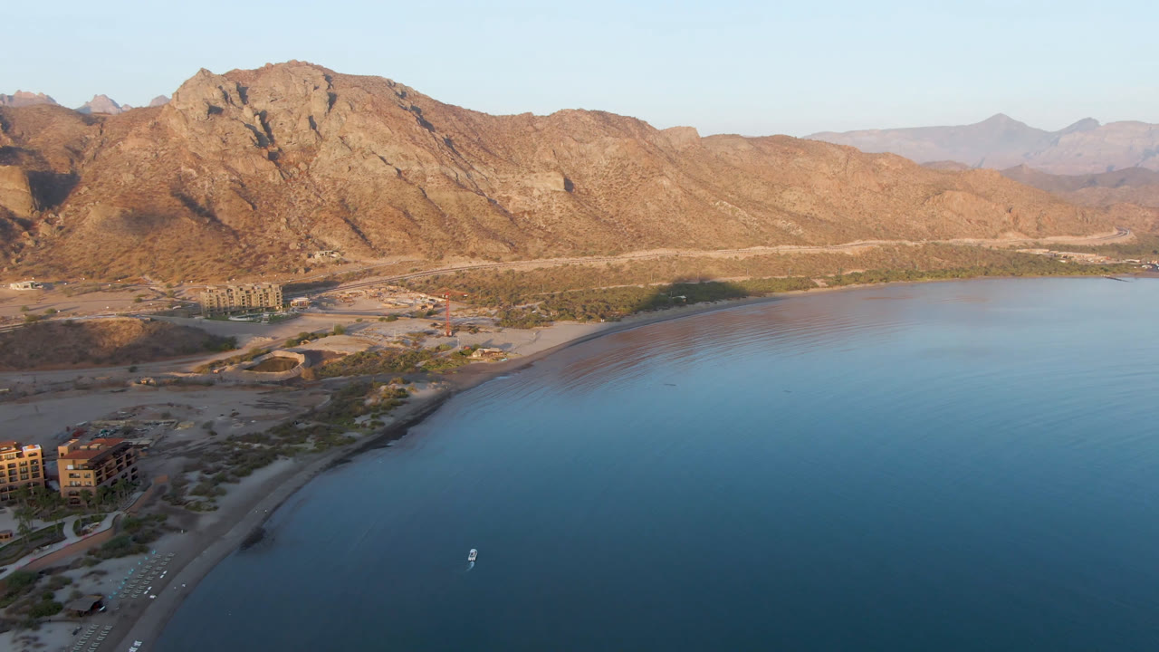 Drone-height view of Danzante Bay showing calm blue water, the beach running diagonally along the left edge, shoreline buildings, distant mountains, and no visible people.