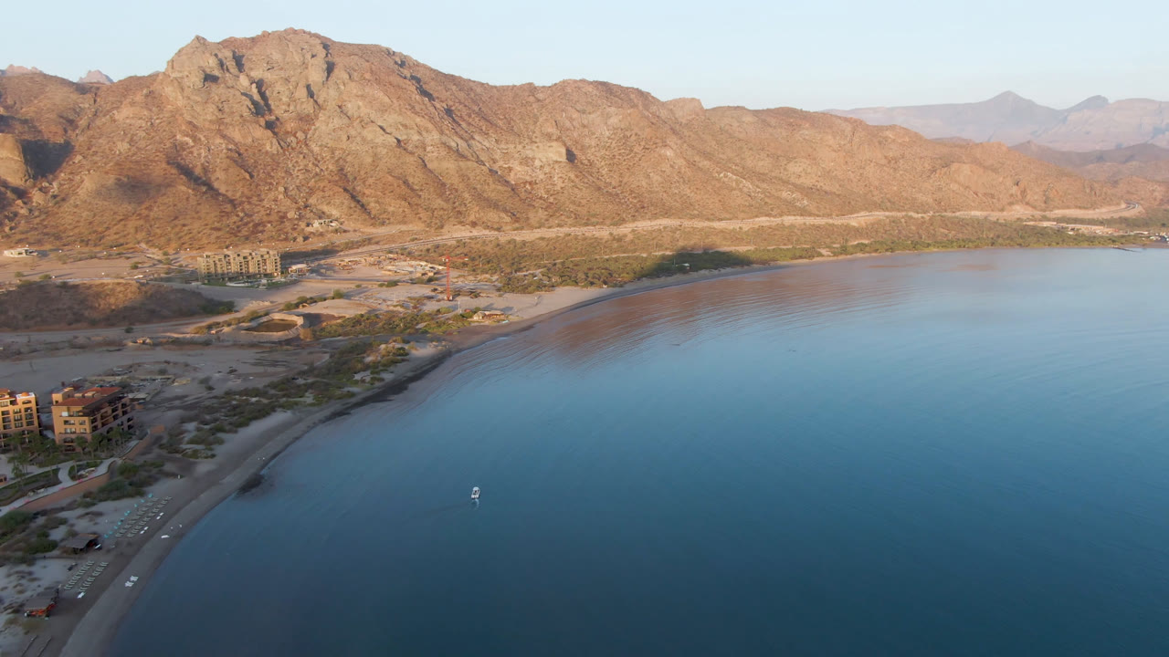 Aerial view of a curved beach, blue bay water, small boat, and mountains.