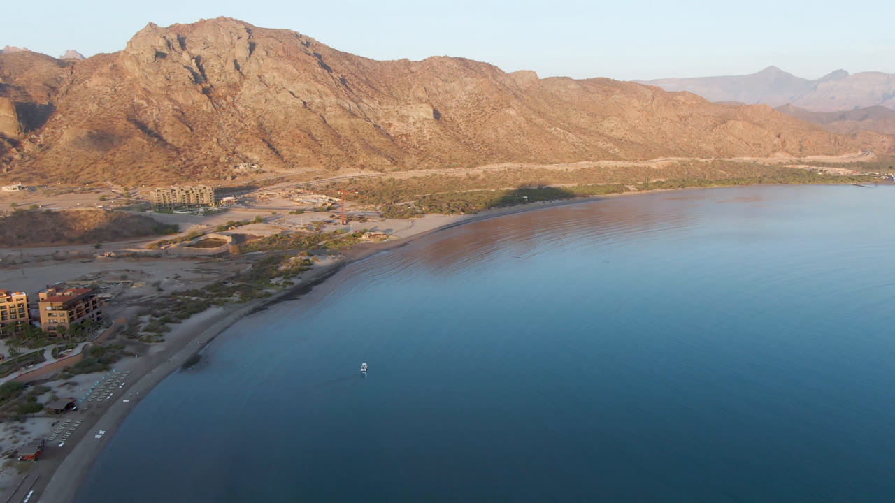 One second into the Danzante Bay Loreto aerial view, shoreline buildings sit near the left edge while the small boat remains offshore on smooth blue water, with no visible people.