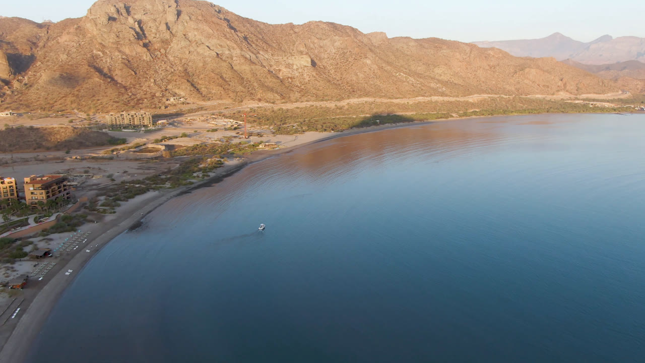 Aerial frame over Danzante Bay Loreto in Baja California Sur, Mexico, showing calm blue water, a curved sandy shoreline, a small boat offshore, coastal buildings, tan mountains, and no visible people.
