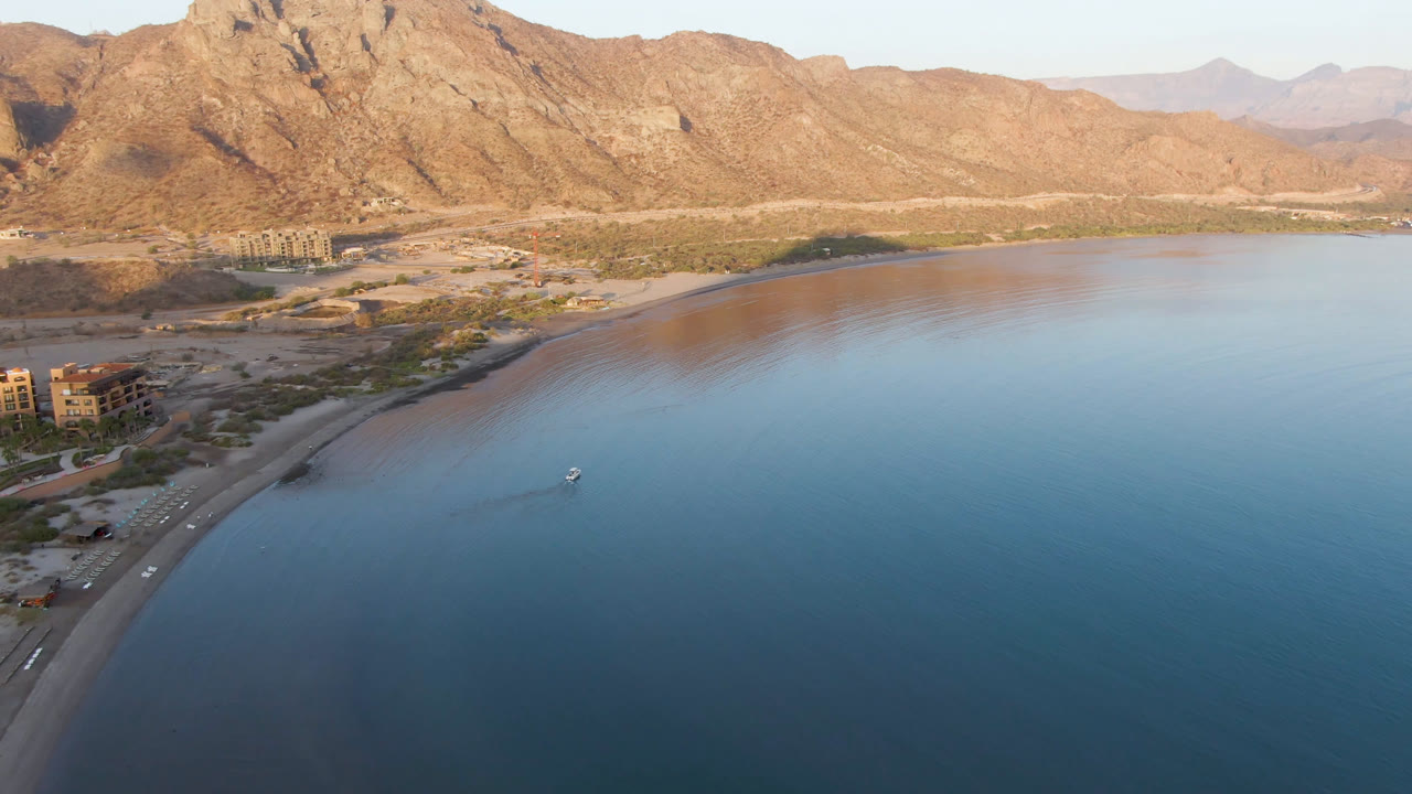 Aerial view of Danzante Bay Loreto with the small boat slightly nearer the center of the water, the beach curving along the lower shore, coastal buildings to the left, mountains behind, and no visible people.