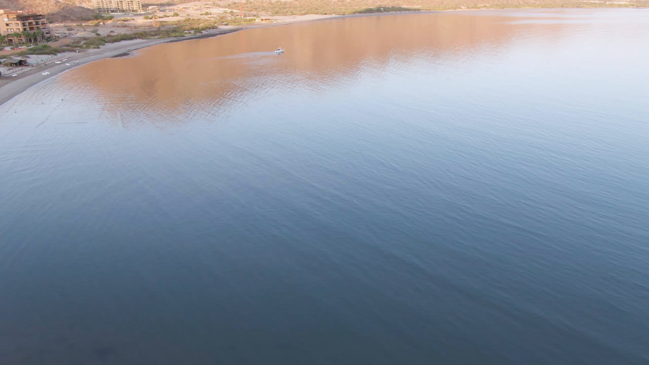 Wide aerial view of calm bay water, a small boat, shoreline buildings, and tan mountains.
