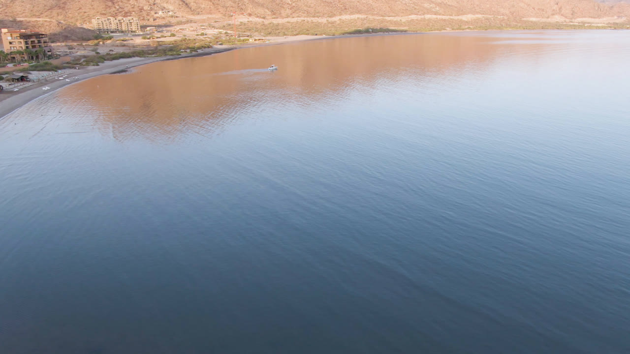 Second frame from the drone view over Danzante Bay, showing the small boat slightly right of the left shoreline, broad smooth water, tan mountain reflection, distant buildings, and no visible people.