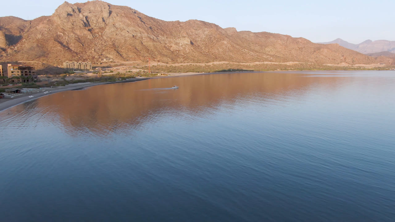 Wide aerial frame of Danzante Bay Loreto with calm blue water in front, brown mountains reflected across the bay, shoreline buildings at the far left, and no visible people.