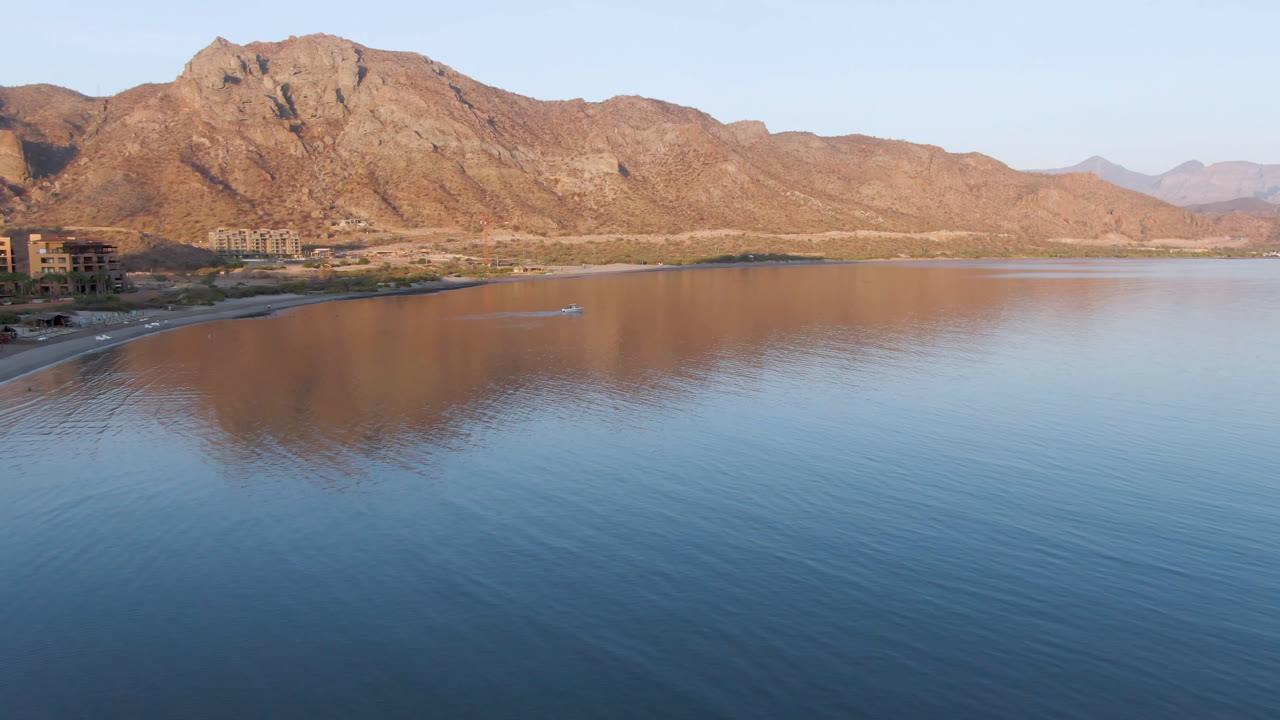 Elevated view over Danzante Bay Loreto with a small boat crossing the reflective water, low shoreline at left, rugged mountains behind it, and no visible people.