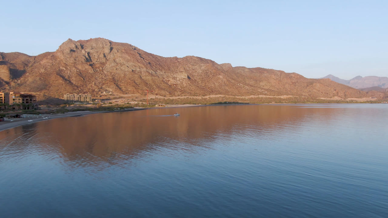 Opening frame of Danzante Bay in Loreto, Baja California Sur, with calm blue water, warm mountains across the horizon, distant shoreline buildings at left, and no visible people.