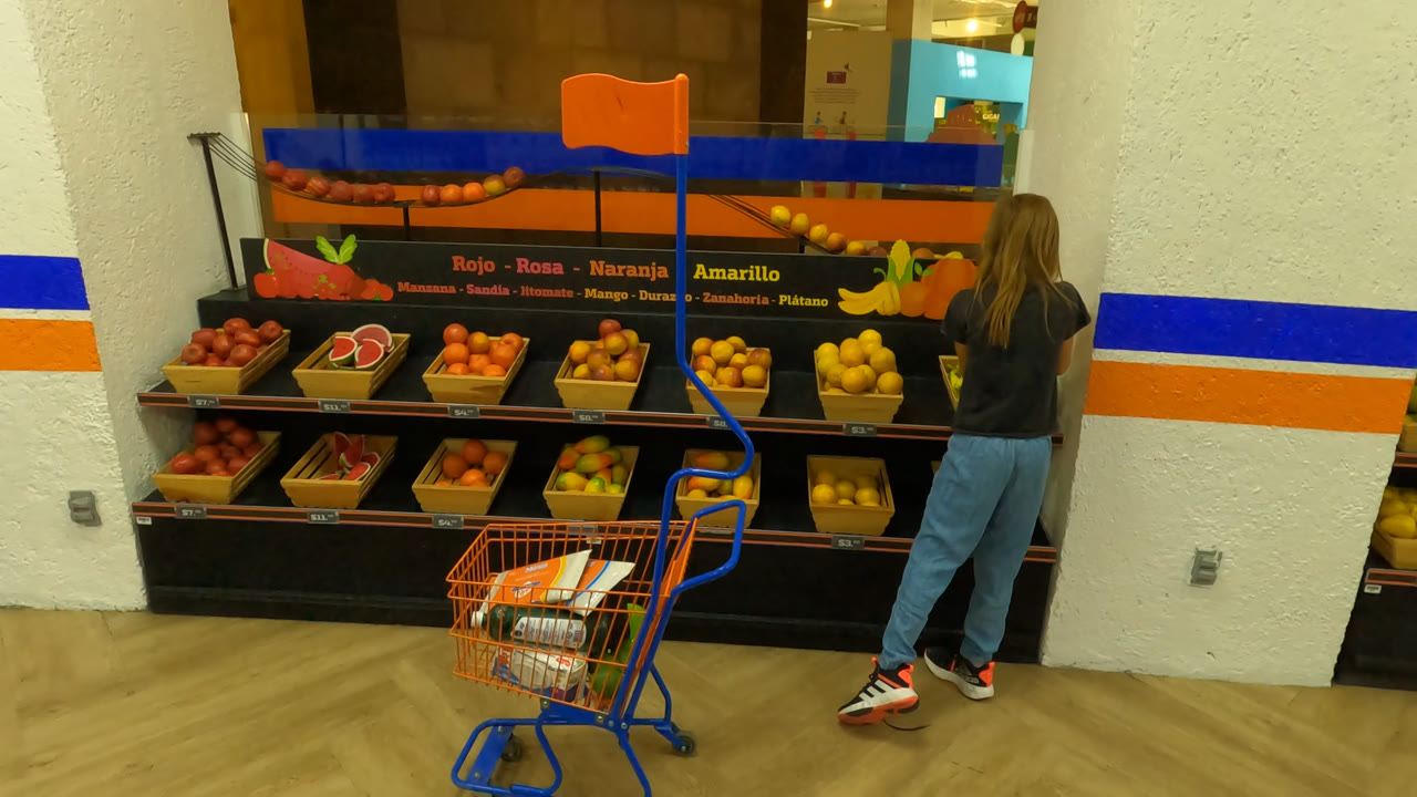 A young girl in Mexico City stands near an orange-and-blue shopping cart while facing a fruit display with bananas, apples, citrus, and watermelon signage.