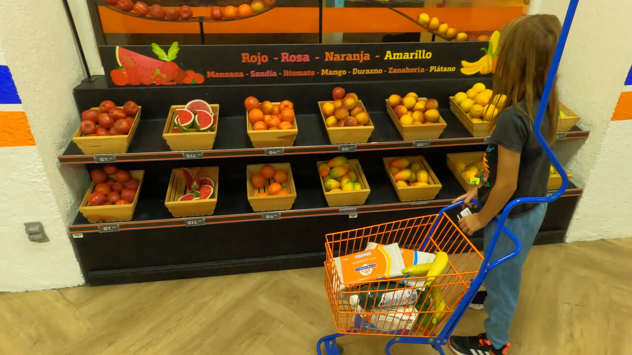 A young girl in Mexico City pushes an orange shopping cart past bins of tomatoes, citrus, and bananas in a brightly lit grocery display.