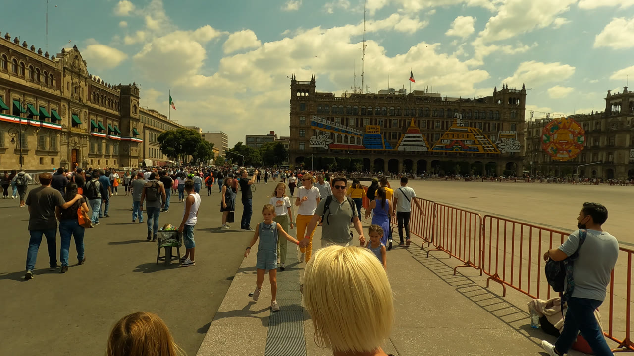 Mexico City plaza scene with a blonde woman seen from behind in the foreground, children and adults walking nearby, red barriers along the right, and large buildings beyond the crowd.