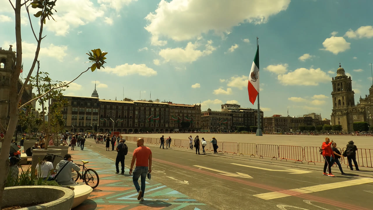 Wide view of a fenced Mexico City plaza with people walking and a Mexican flag.