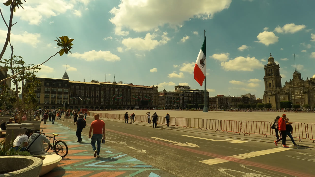 In the next second, pedestrians continue along the edge of the fenced Zocalo Square plaza while the large Mexican flag stands near the cathedral and surrounding buildings.