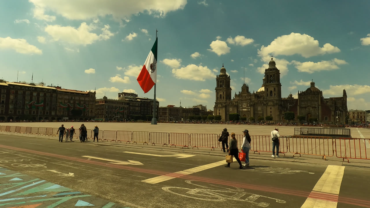 Opening frame of Zocalo Square in Mexico City with the Mexican flag at left, a few pedestrians near metal barriers, road markings in front, and cathedral towers under scattered clouds.