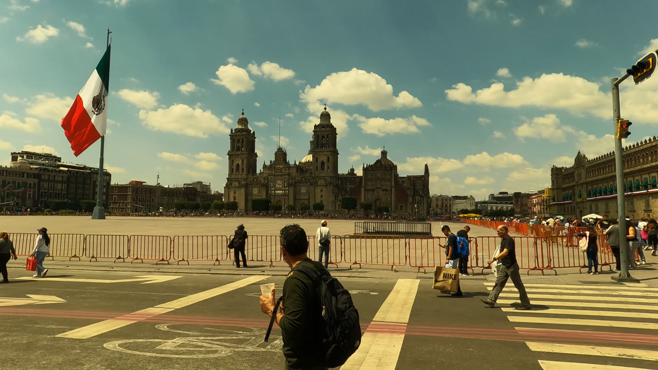 Wide Mexico City view at the start of the clip, showing pedestrians near barricades, a large Mexican flag, striped road markings, and a historic building under scattered clouds.
