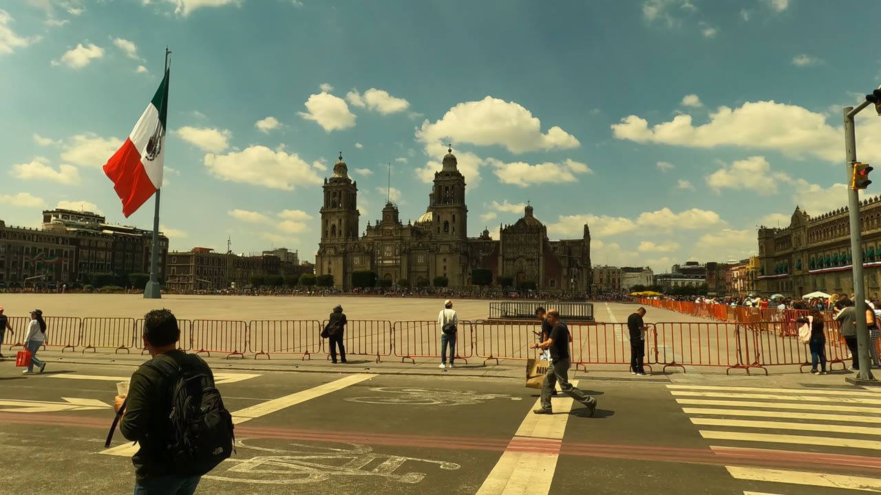Mexico City street frame with a backpacked pedestrian in the foreground, people crossing near orange barricades, and the historic building set behind a large Mexican flag.