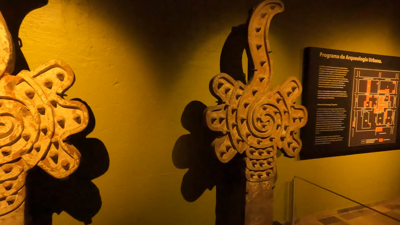 A warm-lit museum wall in Mexico City shows decorative carved forms with spiral relief, an exhibit panel, and part of a glass display case, with no visible people.