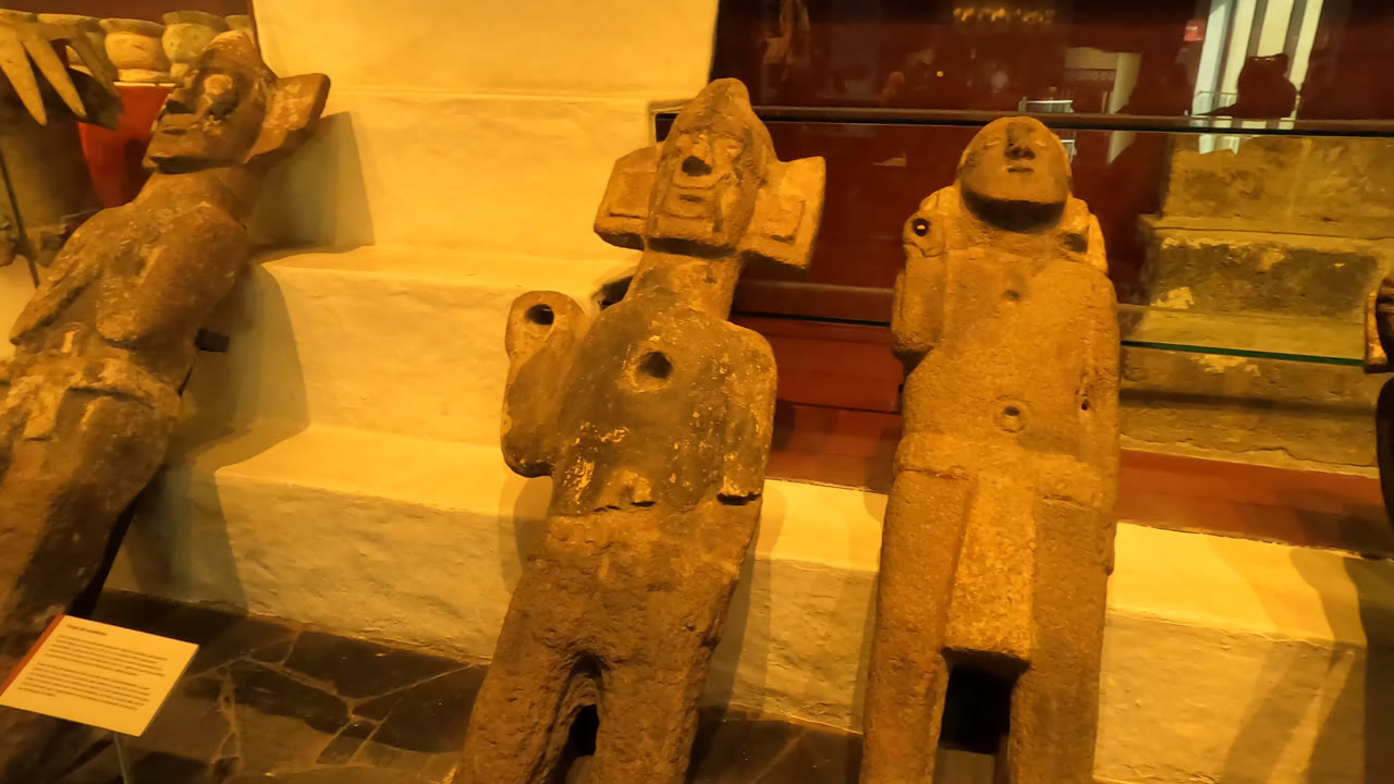Warm-lit stone figures stand in a Mexico City museum display, with a glass case and white wall behind them and no visible people.