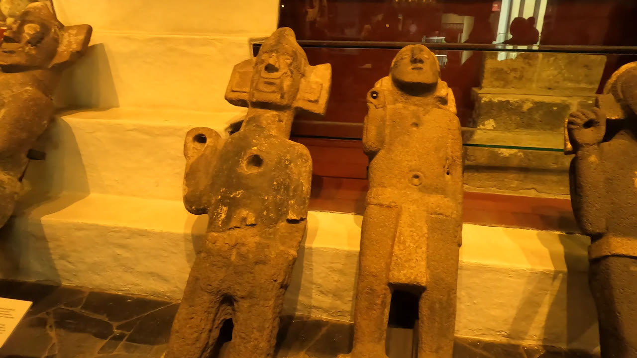 Two central carved stone figures fill the frame in a Mexico City museum, with amber light, a glass display case, and no visible people.