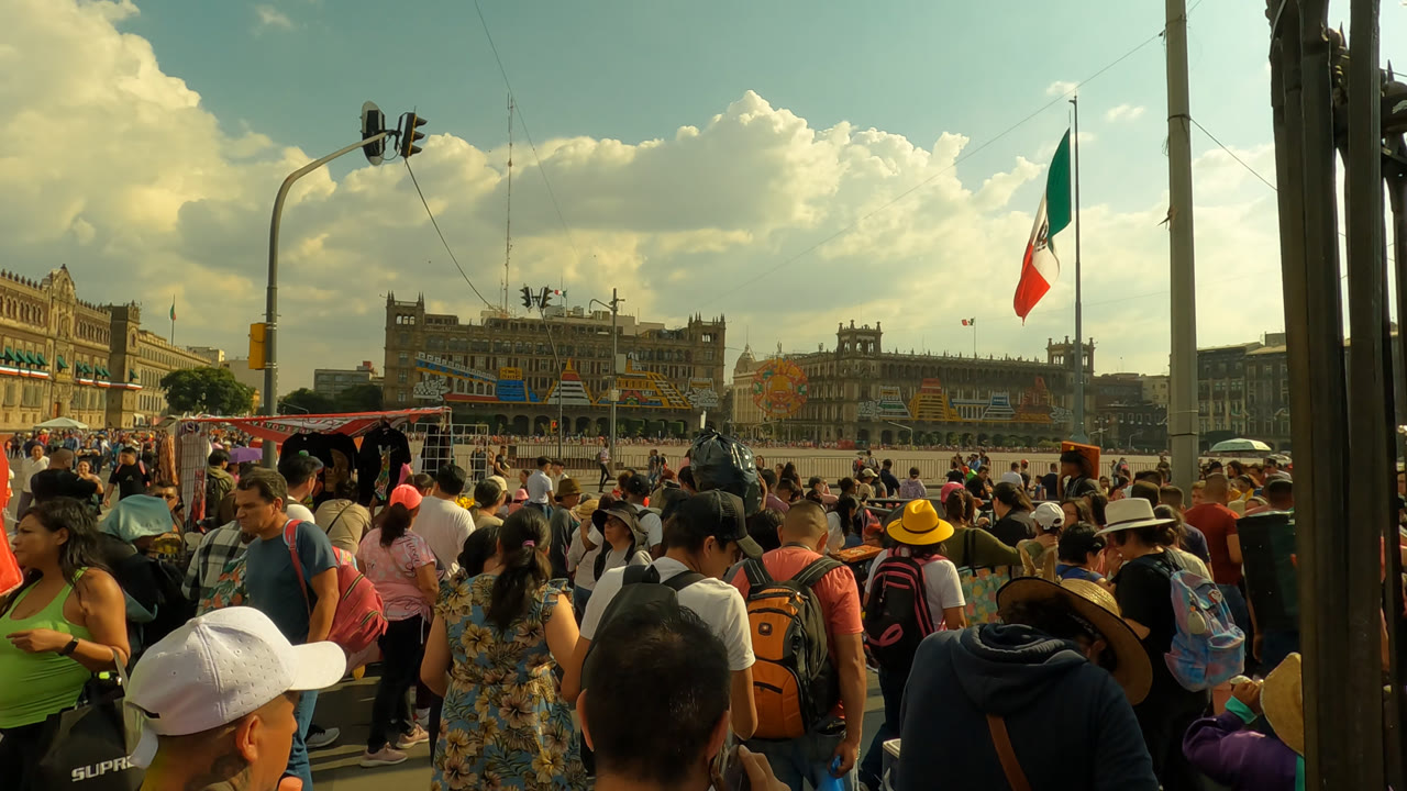 Opening frame of a busy crowd in Zocalo Square, Mexico City, with people in hats and backpacks crossing the foreground and historic buildings under large clouds in the background.
